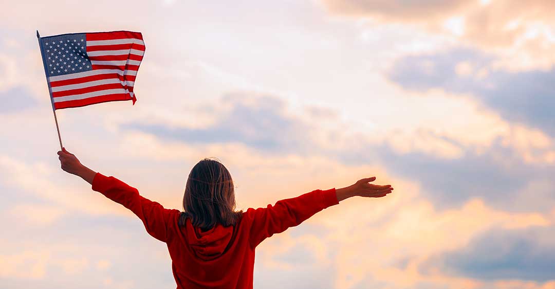 mujer sosteniendo bandera americana mirando al cielo con los brazos abiertos, abogada de la ciudadanía
